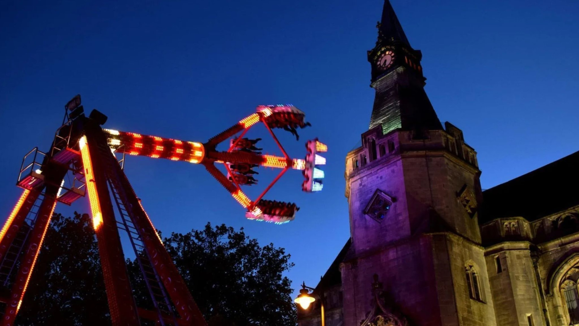 A brightly lit travelling funfair ride spinning beside a historic clock tower at twilight