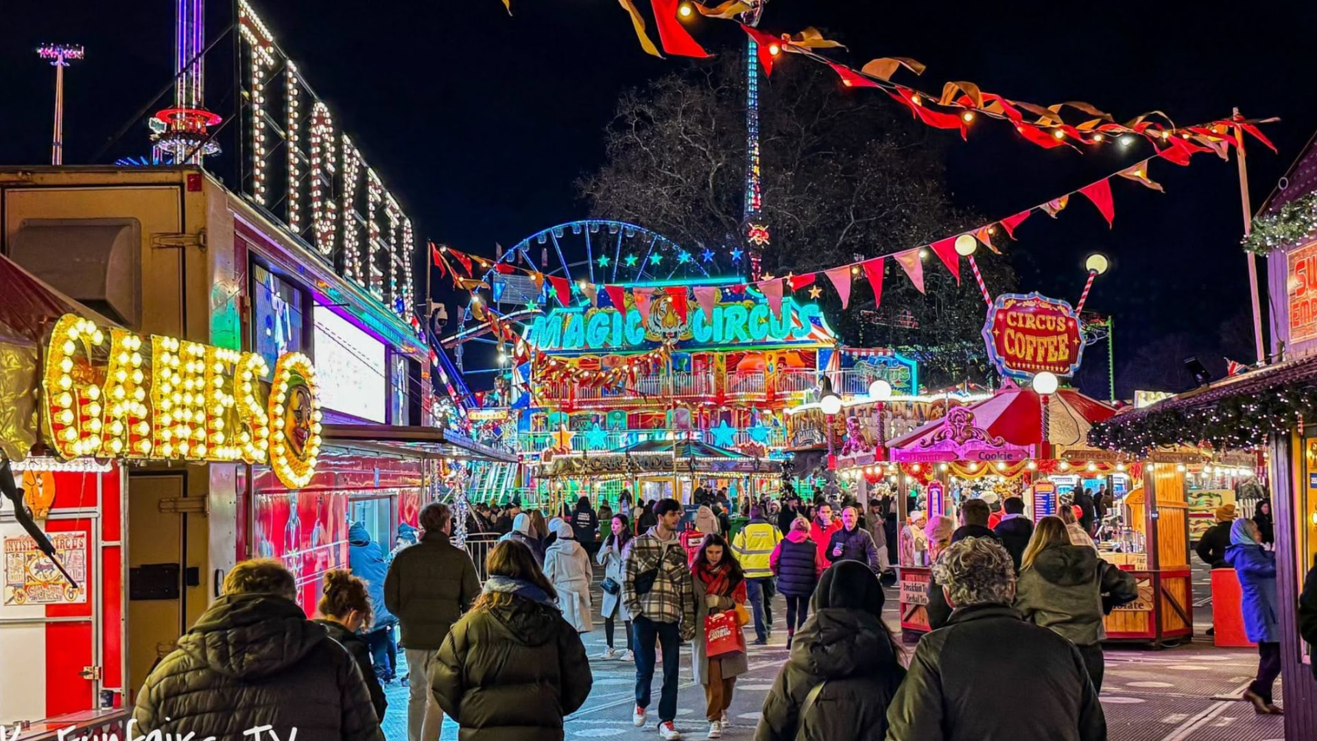 A packed funfair at night, dense crowds walking beneath brightly lit rides